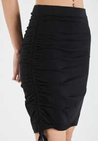 Black fitted skirt made of stretchy fabric, featuring side ruching and ties at the hem for adjustable length and fit.