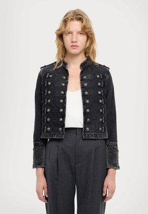 Mladić s plavom kosom do ramena koji nosi crnu vojnu jaknu s gumbima, bijelu majicu i tamno sive hlače.