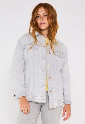 Donna con capelli rossi mossi, che indossa una giacca di denim grigio chiaro e jeans, in piedi contro uno sfondo semplice chiaro.