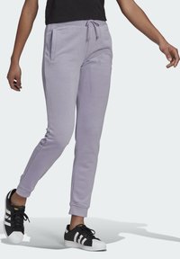 Osoba v levandulových teplácích s tkaničkami a černých teniskách s bílými pruhy, kráčející na pozadí s jednoduchým světlým vzorem.