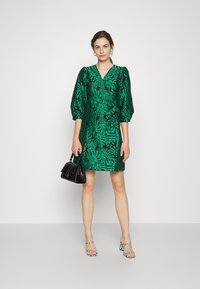 Robe verte avec motif de feuilles, décolleté en V, manches bouffantes et longueur au-dessus du genou. Associée à un sac à main noir et à des sandales à talons noir et blanc.