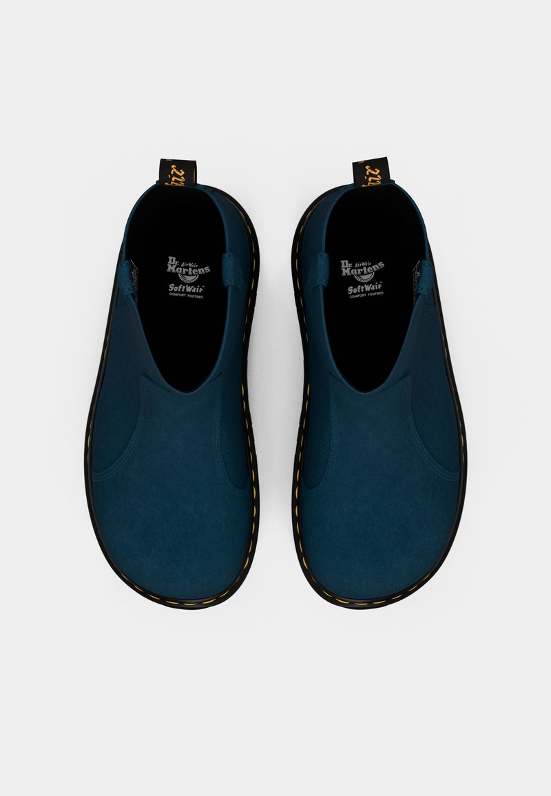 Des chaussures à enfiler bleues avec une texture en daim doux, une semelle en caoutchouc noir et des coutures jaunes. Présentent une languette pour un enfilage facile.