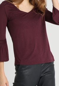 Burgundy V-neck top with three-quarter bell sleeves, textured accents at the shoulders, and a smooth, stretchy fabric. Worn with black pants.