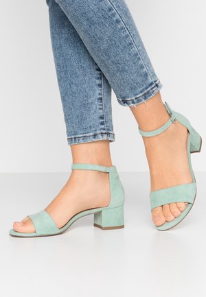 Feet wearing light blue suede block heel sandals with ankle straps paired with cuffed blue denim jeans on a white background.