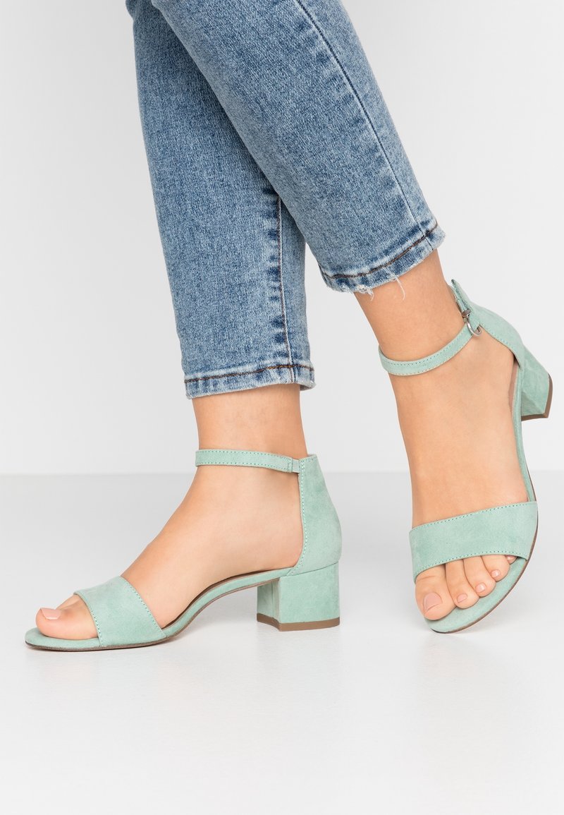 Pieds portant des sandales à talons blocs en daim bleu clair avec des brides autour de la cheville, associés à un jean en denim bleu retroussé, sur un fond blanc.