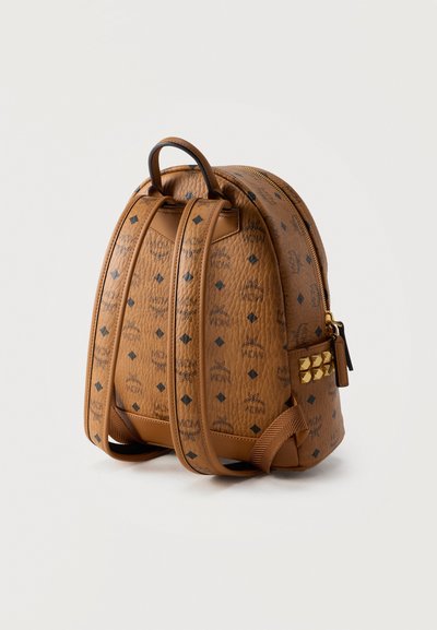 Καφέ δερμάτινο σακίδιο πλάτης με μαύρο σχέδιο λογότυπου, ρυθμιζόμενους ιμάντες, χρυσούς πυραμιδωτούς τρούκς και κλείσιμο με φερμουάρ σε λευκό φόντο.