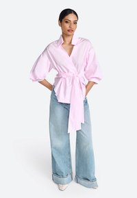 Blusa avvolgente rosa chiaro con scollatura a V profonda, maniche oversize e vita annodata; abbinata a jeans a gamba larga e scarpe a punta.