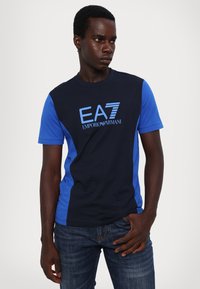 Jūras spēku un zilā kokvilnas T-krekli ar īsām piedurknēm, ar "EA7 Emporio Armani" logotipu gaiši zilā krāsā uz krūtīm. Gluda tekstūra.