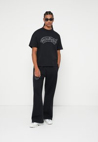 Melnā T-kreklu ar uzrakstu "NAV CITA PLĀNA B" pelēkā krāsā, apvienots ar melnām biksēm. Tam ir brīvais piegriezums un balti apavi.