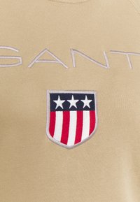 Pullover beige con la scritta "SANT" in ricamo chiaro, dotato di una toppa a forma di scudo con tre stelle bianche su strisce rosse e bianche.
