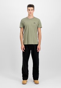 Olivgrünes Baumwoll-T-Shirt mit Rundhalsausschnitt, kurzen Ärmeln und einem kleinen schwarzen Logo auf der Brust. Kombiniert mit schwarzen Hosen und beigen Schuhen.