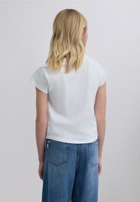 Weißes Kurzarmshirt mit Rundhalsausschnitt, das eine lockere Passform bietet. Kombiniert mit blauen hoch taillierten Jeans, die ein schlichtes Rückendesign zeigen.