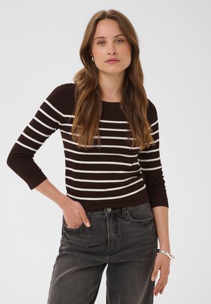 Mujer con cabello largo y castaño, vistiendo un suéter negro con rayas horizontales blancas y vaqueros gris oscuro, posando con una mano en el bolsillo.