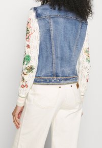 Denimová vesta s krajkovými rukávy, zdobená květinovou výšivkou, vrstveným designem a bílými textilními akcenty. Pohled zezadu ukazuje detaily a švy.