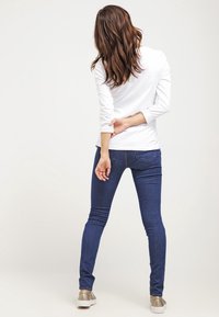 Kvinna med brunt hår står vänd bort, klädd i en vit långärmad tröja, blå jeans och beige sneakers mot en neutral bakgrund.