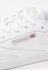 Valkoinen nahkakengät, joissa on teksturoitu pohja, Reebok-logo, hopeiset yksityiskohdat ja Union Jack -yksityiskohta logon lähellä. Klassinen muotoilu.