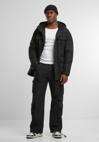 Sort hættetrøje med flere lommer, hvid t-shirt og sorte cargo bukser. Læder sneakers med grønne og hvide detaljer.