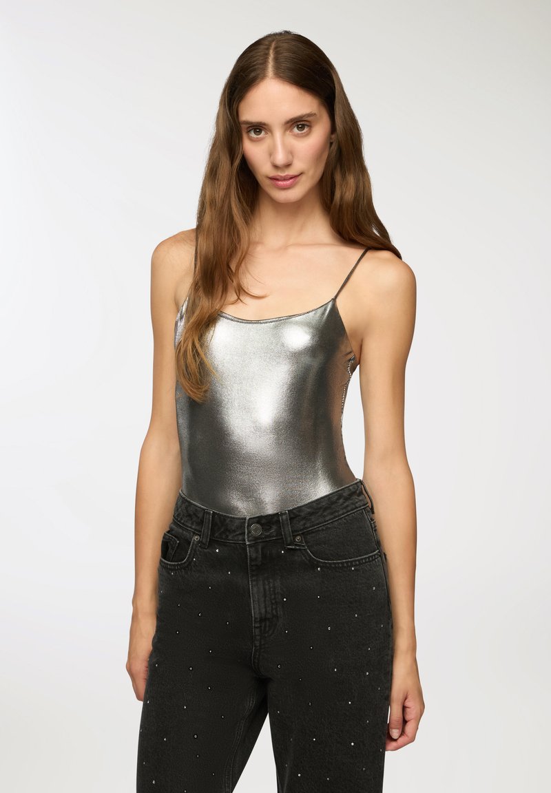 Metaliczny srebrny kostium z cienkimi ramiączkami w połączeniu z czarnymi dżinsami ozdobionymi rozproszonymi cyrkoniami. Błyszcząca tekstura i dopasowany krój.