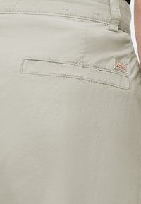 Gros plan de la poche arrière d’un pantalon beige avec une fente horizontale et une petite étiquette rectangulaire de la marque BOSS au-dessus de la poche.
