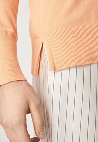 Jasnopomarańczowy sweter z dzianiny z prążkowanymi mankietami i bocznymi rozporkami u dołu, zestawiony w paski. Skup się na miękkości i kontrastach kolorystycznych.