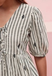 Robe rayée en crème et noir avec des manches bouffantes, un devant à lacets et des détails de broderie florale. Tissu doux et texturé.