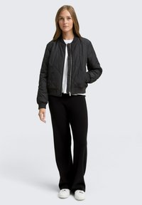 Donna che indossa una giacca bomber trapuntata nera, una camicia bianca, pantaloni larghi neri e sneakers bianche, in piedi di fronte a uno sfondo chiaro e semplice.