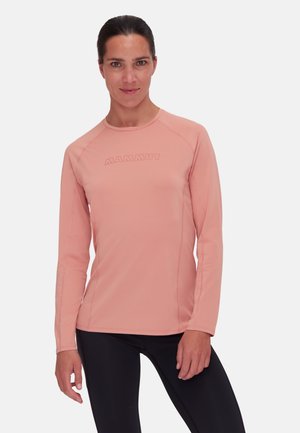 Kvinde iført en tætsiddende langærmet lyserød Mammut trøje og sorte leggings, stående foran en ensartet lys grå baggrund.