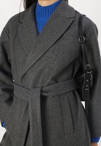 Grijze wollen wikkeljas met een ceintuur in de taille, voorzien van revers met insnijding en zijzakken, gedragen over een felblauwe gebreide trui. Inclusief zwarte tasdetails.