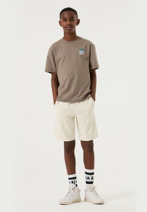 Jugendlicher Junge steht mit Händen in den Taschen, trägt ein braunes T-Shirt, beige Shorts, weiße High-Top-Sneaker und weiße Socken mit schwarzem Schriftzug.