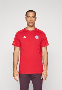 Rød treningst-skjorte laget av myk stoff. Har hvit Adidas-logo og Bayern München-emblem på brystet, med oransje striper på skuldrene.