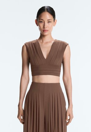 Žena nosiaca hnedý crop top bez rukávov s plisovaným dizajnom a zodpovedajúce vysoký pás plisovaných nohavíc, stojaca pred jednoduchým svetlým pozadím.