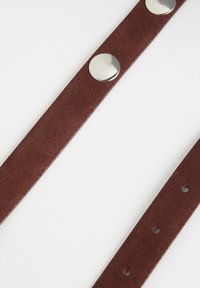 Sangle en suède marron avec des boutons métalliques et plusieurs trous pour les réglages. Texture lisse avec une forme plate et allongée.