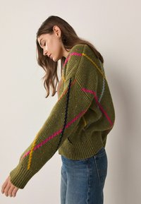 Green knitted sweater with ribbed texture, featuring pink, yellow, blue, and black diagonal stripes. Long sleeves and a relaxed fit.