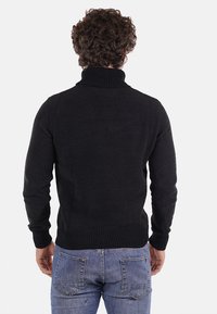 Maglione nero lavorato a maglia con collo alto, polsini e orlo a coste. La texture è spessa e accogliente. Indossato da una persona vista di spalle.