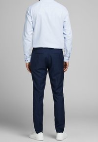 Man står vänd bort med en ljusblå skjorta, marinblå kostymbyxor och vita sneakers mot en neutral grå bakgrund.