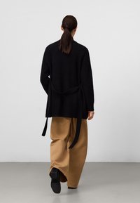 Kvinna med mörkt hår i hästsvans som bär en svart tröja med hängande remmar och vida tanfärgade byxor, går på ett grått golv mot en vit vägg.