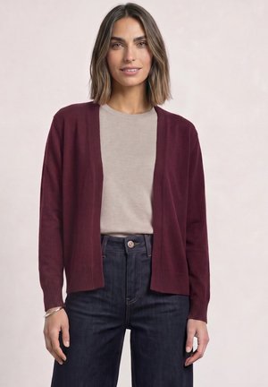 Kvinde iført en mørkerød åben-front cardigan over en lys beige top og mørkeblå jeans, stående foran en ensfarvet lys baggrund.