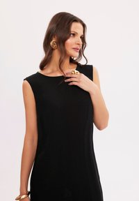 Czarna sukienka bez rękawów o luźnym kroju. Gładki materiał i okrągły dekolt. Zawiera złote akcesoria: duże kolczyki i pierścionek.