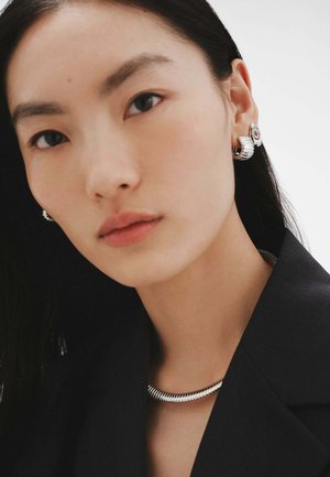 La joyería de plata presenta un collar texturizado y unos pendientes a juego con un diseño ondulado. El sujeto lleva puesto un blazer negro y tiene el cabello lacio.