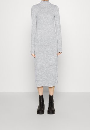 Femme portant une robe midi tricotée à manches longues, gris clair, associée à des bottines noires, debout devant un mur clair uni.