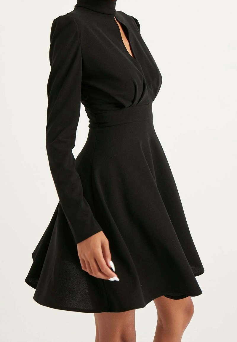Femme portant une robe noire ajustée à manches longues, col montant et jupe évasée sur un fond uni.