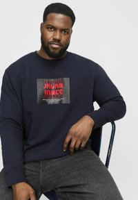 Námornícka mikina vyrobená z bavlny s rebrovanými manžetami, s veľkým grafickým potlačou v červenej a čiernej farbe s nápisom "JACK & JONES" na prednej strane.