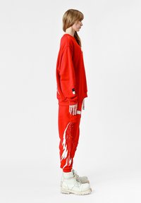 Sudadera roja de gran tamaño con tela texturizada, combinada con pantalones a juego que presentan un diseño de plumas blancas y botas de combate blancas con cordones.