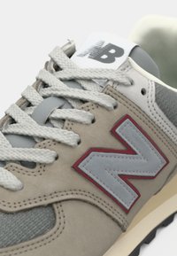 Sneaker in suede beige e tessuto con accenti grigi e un logo rosso. Punta rotonda, lacci bianchi spessi e suola in gomma leggera.