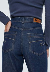 Jean en denim bleu foncé avec des coutures jaunes, poches arrière carrées et une étiquette de marque en cuir brun sur la ceinture, portés avec un haut bleu marine.