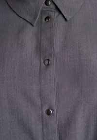 Pelēka krekla ar apkakli, kuram ir vertikāla rinda ar četriem tumšiem pogu spiedpogu slēdzējiem uz teksturētas auduma virsmas.