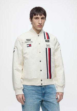 Ung mand iført en hvid racerjakke med Cadillac-, Tommy Hilfiger- og GM-logoer, kombineret med lyseblå jeans, på en ensfarvet hvid baggrund.