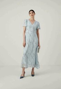 Žena stoji u svijetloplavoj haljini do gležnjeva s lepršavim rukavima, držeći clutch torbicu i noseći tamne cipele na visoku petu.