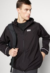Schwarze Windjacke mit Kapuze, Reißverschluss, elastischen Bündchen und einem Logo auf der Brust. Verfügt über reflektierende Akzente an den Ärmeln.