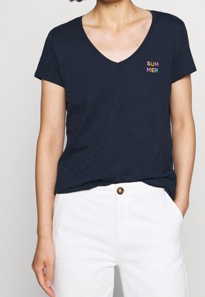 Kvinde iført en mørkeblå V-hals T-shirt med farverig "SUMMER" tekst på venstre bryst og hvide bukser med hånden i lommen.